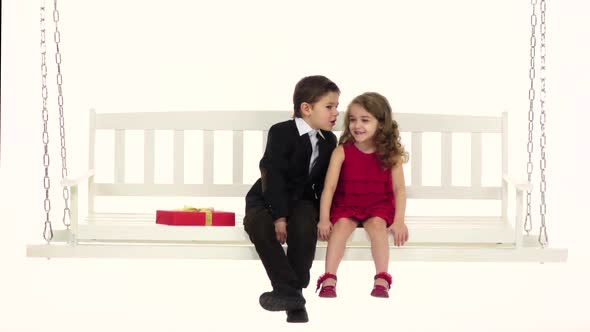 Children Ride on a White Swing Tell Each Other Secrets in the Ear. White Background. Slow Motion alt