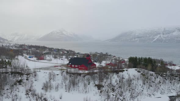 Aerial drone orbits around Kåfjord Church in Norway. Snow covered coastline, with mountainous backgr