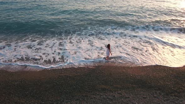 Aerial View of Excited Young Woman Walking in Foamy Mediterranean Sea Waves on Beach at Sunset