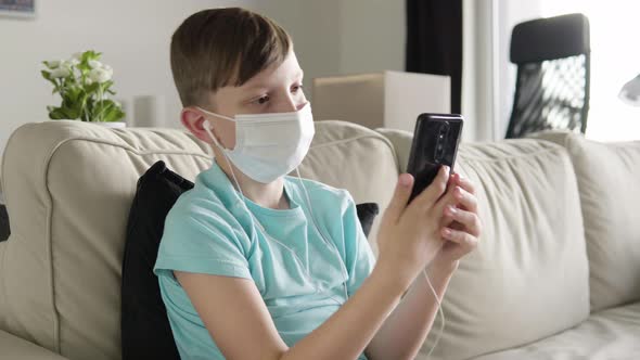 A Young Boy in a Face Mask Works on a Smartphone and Listens To Music with Earphones