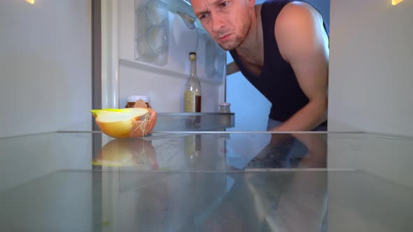 man looks into refrigerator, sniffs an onion and takes a bottle of alcohol.