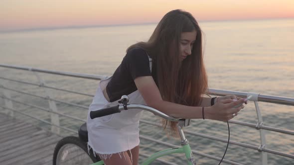 Young Attractive Woman Uses a Smartphone and Riding Vintage Bike Near the Sea During Sunrise or