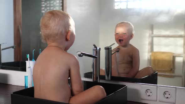 Cute Little Caucasian Baby Sitting in the Sink Playing with Water Looking in the Mirror