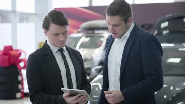 Portrait of Male Caucasian Car Dealer and Client Using Tablet and Looking Around in Car Showroom