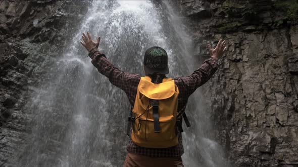 Traveler Man with a Yellow Backpack Standing on the Background of a Waterfall