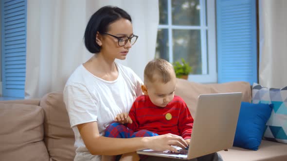 Young Woman Sitting on Couch with Baby Checking Financial Report on Laptop Working From Home