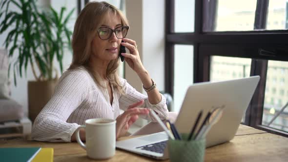 Businesswoman Talking on Phone and Sitting at Table with Laptop in Home Office