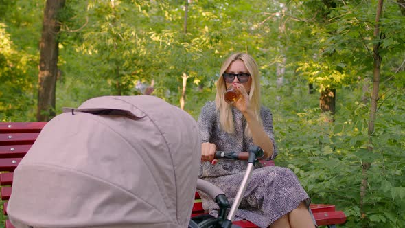 A Young Mother in Sunglasses Drinks Beer on a Walk with a Stroller. An Irresponsible Mother Drinks