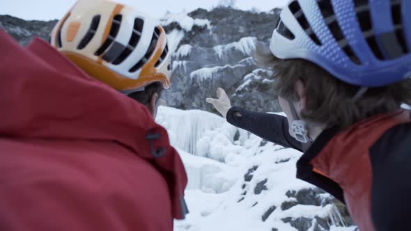 Two men point and discuss a climbing route for ice climbing on a frozen waterfall in the mountains.