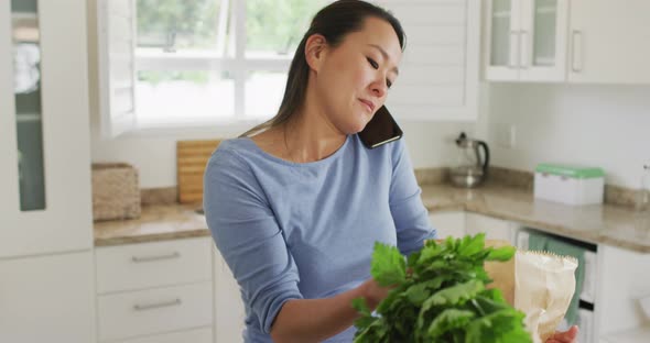 Happy asian woman unpacking groceries and using smartphone in kitchen