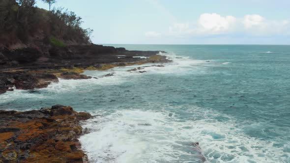 Drone camera moves over the foaming waves hitting rocky shore on a clear day (Kauai, Hawaii, USA)