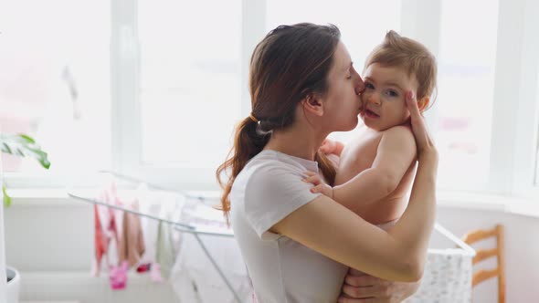 Satisfied Young Mom Kisses and Hugs Her Baby at Big Window at Home