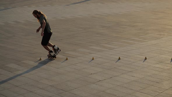 Young Long-haired Man Roller Skater Is Dancing Between Cones in a Nice Evening in a City Park