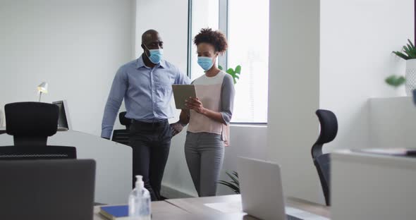 Diverse businessman and businesswoman in face masks, woman holding tablet in office