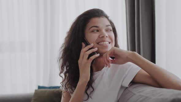 Cheerful Black Girl Talking On Mobile Phone Sitting On Couch At Home And Playing With Hair Free