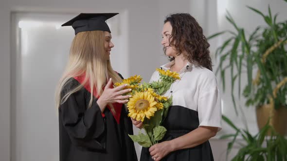 Happy Proud Mother and Daughter Graduate with Bouquet of Flowers Standing Indoors Smiling Looking at