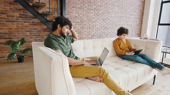 Father and Son Use Gadgets Sitting on White Couch at Home