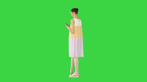 Young Beautiful Girl in Whiteyellow Dress Stands Looking at Her Smartphone and Smiles on a Green