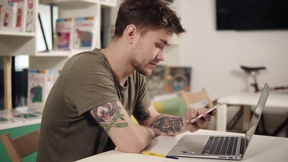 Attractive Programmer with Tattooes on His Arms Scrolling Something on His Smartphone While Sitting