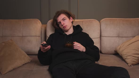 Bored Young Man Watch TV on Couch at Home Drink Coffee or Tea