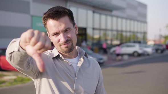 A Young Caucasian Man Shows a Thumb Down to the Camera and Shakes His Head in an Urban Area