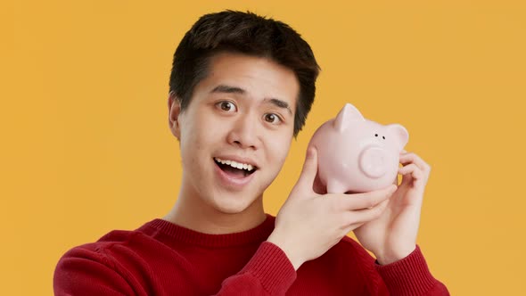 Rich Asian Man Shaking Piggybank Full Of Money Yellow Background