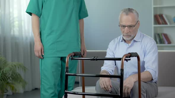 Old depressed male sitting on couch and looking on walking frame, nursing home