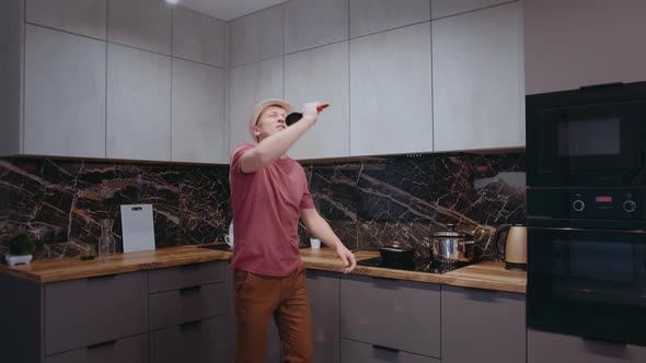 Young Man in a Hat Dancing and Singing with a Ladle in His Hand in the Kitchen While Cooking