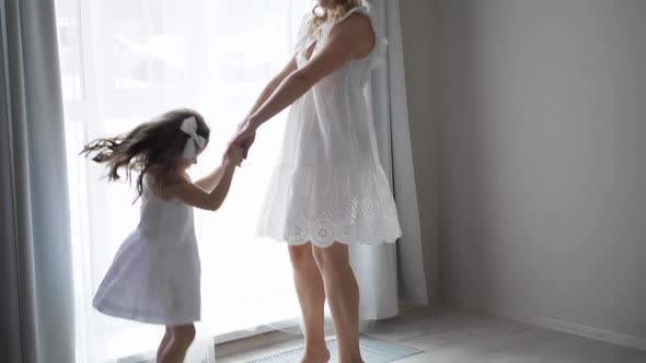 Beautiful Family Mother and Daughter in White Dresses are Sitting on the Floor By Large Window