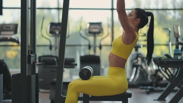 Hispanic Woman Sitting on a Simulator in the Gym Pulls a Metal Rope with the Weight Pumps Up the