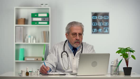 Aged Male Doctor Reviews the Patient's Analyzes on a Laptop and Makes Notes in His Medical History
