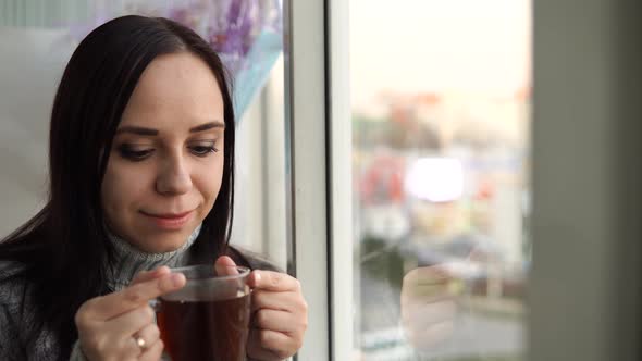 A Young Beautiful Woman in a Gray Sweater Drinks a Tea Looking Out the Window on the Balcony