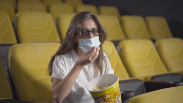 Portrait of Absorbed Young Woman in Face Mask Trying To Eat Popcorn in Cinema. Engrossed Caucasian