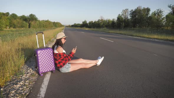 Traveler Woman with Suitcase Sits on Road and Waiting for a Car To Travel. Pretty Young Woman