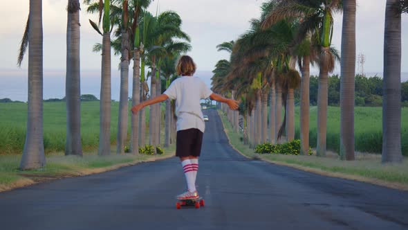 A Teenager with Long Hair Rides a Skateboard Along a Beautiful Road with Palm Trees