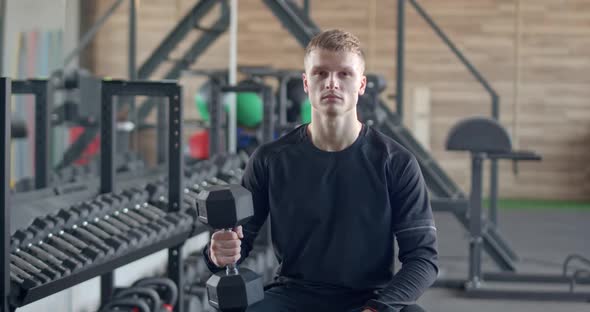 Young Man in the Gym Sitting on a Bench and Holding a Dumbbell in His Hand a Guy Looks at the Camera