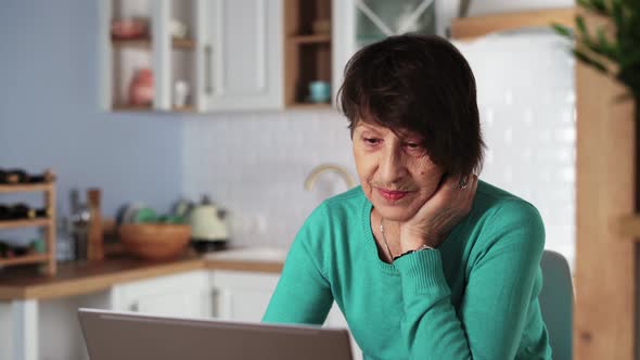 Elderly Woman Laughing and Smiling Using Laptop at Home in Kitchen