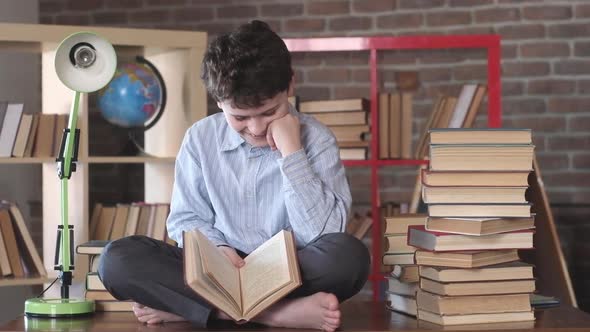 Schoolboy sits on a Desk at home. He holds an open book in his hands and laughs