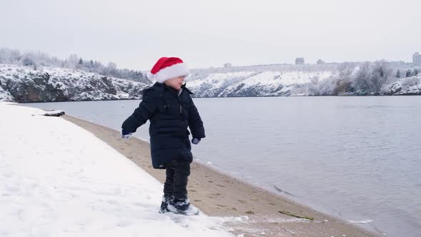 A Beautiful Christmas Child in a Santa Claus Hat Plays on the Bank of the River