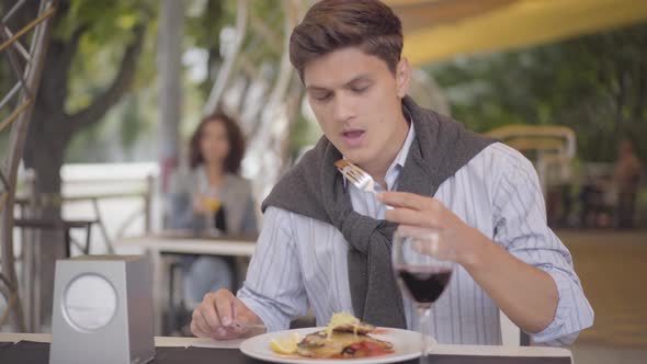 Young Man Eating Tasty Meat Food at Street Cafe and Drinking Red Wine