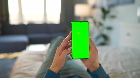 Woman at Home Lying on a Bed and Using Smartphone with Green Mockup Screen in Vertical Mode