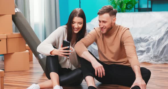 Happy Young Caucasian Couple Use Digital Smart Phone Discuss Interior Design Renovation Ideas Sit on