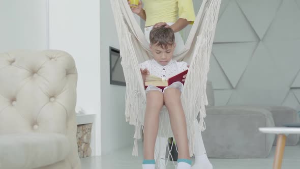 Wide Shot Portrait of Absorbed African American Little Boy Sitting on Hanging Chair and Reading Book