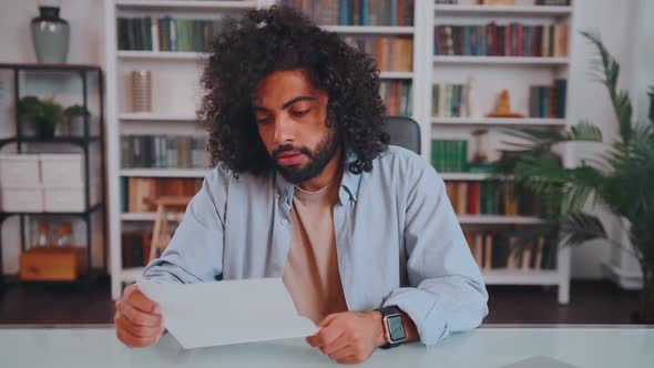 Young Arabian Man Entrepreneur Holds Sheet of Paper Sits at Table with Laptop