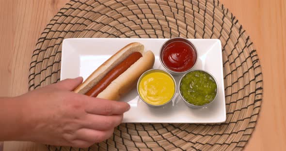 Top down view of a hand placing hot dogs on a plate beside ketchup, mustard, and relish.