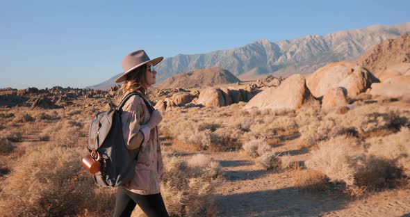 Smiling Fashionable Young Woman in Hat Walking By Desert Landscape at Sunrise