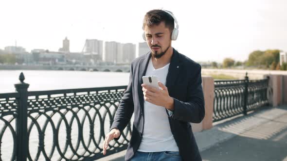 Joyful Man Walking Along River Embankment Dancing Wearing Headphones and Looking at Smartphone