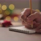 Slow Motion Orbit Shot Man Writes Christmas Gifts Notepad Pencil Under Warm Light Evening - VideoHive Item for Sale