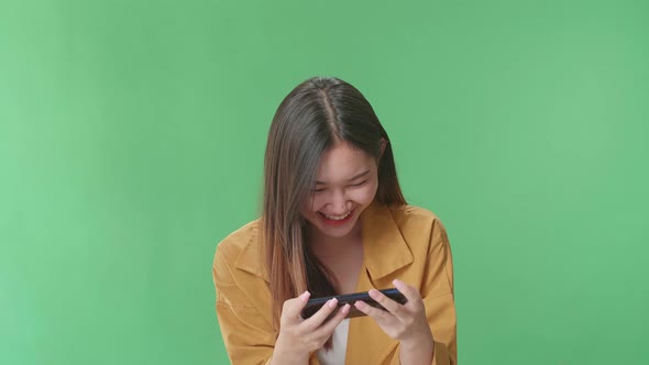 Laughing Asian Woman Looking At The Phone Screen While Standing On Green Screen In The Studio