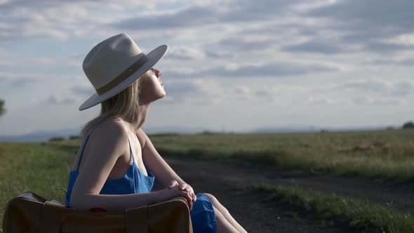 Girl in blue dress with suitcase sit on country road in summer.